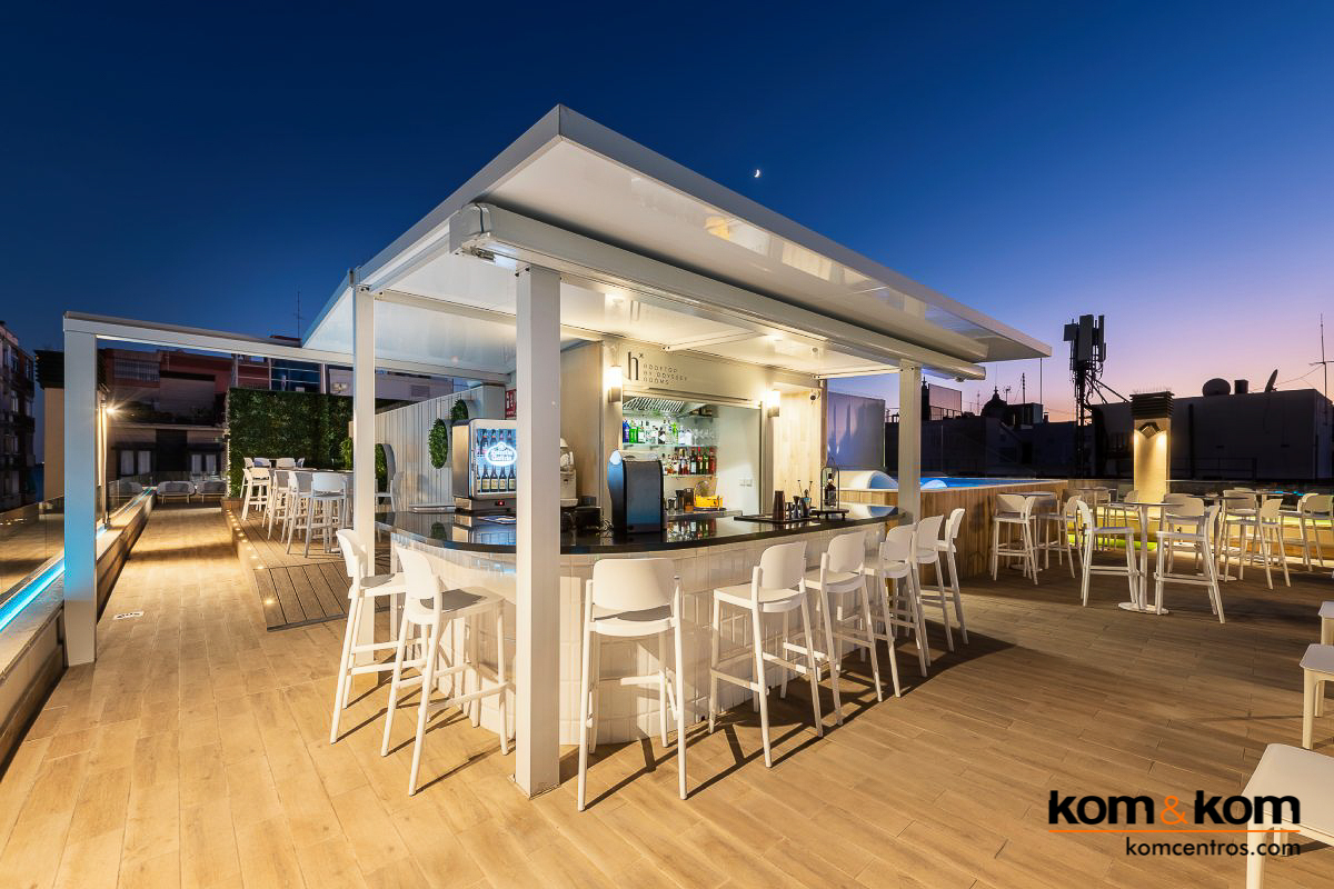 Bar de terraza en azotea de hotel con encimera de acero inoxidable, estantes de cristal con botellas de c&oacute;ctel variadas, cocteleras y fregadero. Dise&ntilde;o elegante y profesional para bar de exteriores.